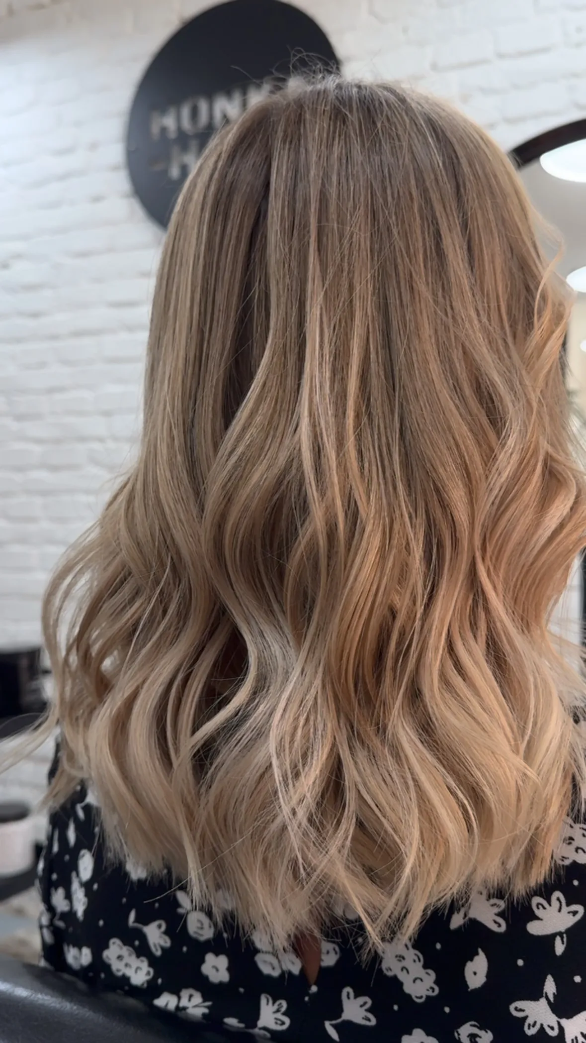 Dlouhé blond vlny s balayage přechodem, pohled zezadu proti bílé cihlové stěně